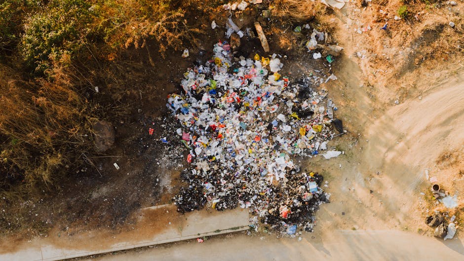 An aerial view of a large heap of mixed domestic rubbish scattered on an uneven patch of ground adjacent to a paved pathway. The waste includes various plastic bottles, food wrappers, and paper packaging, with some items partially buried in dark soil and others resting on a light brown, dusty surface. Surrounding the rubbish, there are patches of dry, sparse vegetation and a section of exposed earth, indicating an outdoor area, possibly a site of illegal dumping or an unofficial waste disposal spot. On the right side of the image, a section of a circular manhole cover or pipe is visible, along with evidence of recent disturbance in the soil from ground leveling or excavation. This scene exemplifies unmanaged waste accumulation in a semi-rural or suburban environment, highlighting the importance of proper rubbish removal services such as those offered by House Clearance Enfield for professional and environmentally responsible waste handling.