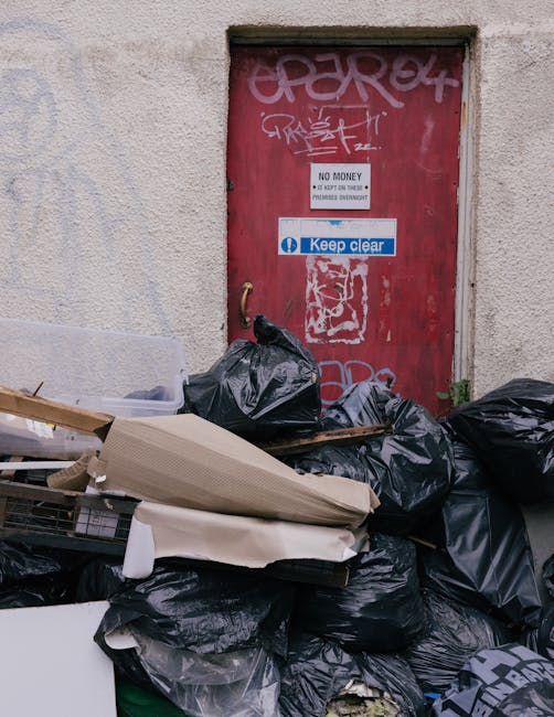 Piles of black and grey rubbish bags overflowing onto the pavement in front of a red metal door on a beige-textured building wall. The bags are filled with waste, some appearing torn or bulging, and are stacked haphazardly, with a cardboard box and wooden pallet leaning among them. The door features graffiti tags in white and pink spray paint, with several signs attached, including one stating 'NO MONEY' and another blue sticker reading 'Keep clear.' The scene appears to be an external urban setting, illustrating a cluttered area awaiting waste collection or disposal, associated with private rubbish removal services such as those offered by House Clearance Enfield, highlighting the importance of proper rubbish management in local environments.