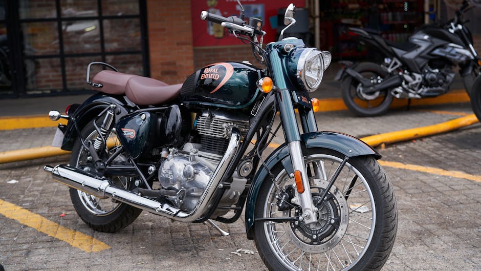 A dark-colored Royal Enfield motorcycle with a tan leather seat parked in a designated parking space on a paved surface. The motorcycle features a large front headlamp, orange turn signals, and a chrome exhaust pipe running along the right side. The fuel tank is adorned with a Royal Enfield logo and has a glossy finish, reflecting nearby light. The wheels have metal spokes and black tires with visible tread patterns. In the background, another motorcycle is partially visible to the right, parked parallel to the first, and a brick building with glass windows and red framing can be seen behind them. The parking area is bordered by a low yellow curb, and the setting appears to be outside an urban or commercial building, with natural lighting casting soft shadows. The scene suggests a casual parking spot where motorcycles are stored, potentially associated with a service or storage area related to motor vehicle or waste handling, as part of a broader context of local transportation or logistical support services.
