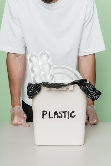 A person wearing a white T-shirt and transparent disposable gloves stands behind a white plastic bin labeled 'PLASTIC'. The bin contains a crumpled black plastic garbage bag. A clear plastic egg carton with multiple depressions for eggs is placed on top of the bin, partially leaning against the black bag. The background features a plain light green wall, and the scene is lit with soft, even lighting, emphasizing the focus on waste items designated for plastic recycling or disposal, representative of rubbish collection and waste management services. The image visually highlights the sorting and handling of plastic waste, relevant to private rubbish collection and on-site clearance services offered by companies like House Clearance Enfield.