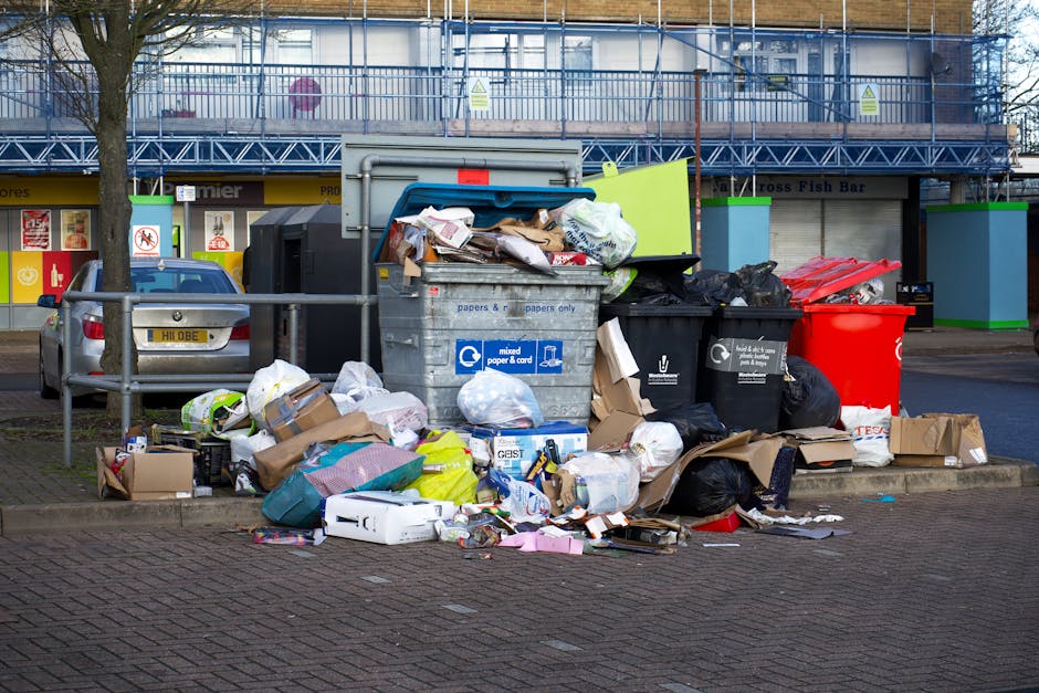 A collection of overflowing waste and rubbish bags situated on a paved sidewalk adjacent to a curb in an urban area. The scene includes black, red, and grey wheelie bins, with some lids open and debris spilling out, containing mixed waste such as cardboard boxes, plastic bags, paper, and packaging materials. Surrounding the bins are scattered boxes, paper, and discarded items, some flattened or torn, contributing to an unkempt appearance. Behind the rubbish, there are parked cars, including a silver hatchback, and a building with storefronts painted in yellow, green, and blue, featuring signage for local businesses. A metal railing separates the rubbish area from the parking and sidewalk, and a large tree trunk is visible on the left. The background features a multi-story building with scaffolding and safety barriers, indicating construction or refurbishment work nearby. The lighting suggests a cloudy day, emphasizing the cluttered, unmanaged waste scene typical of private rubbish collection in an urban environment.