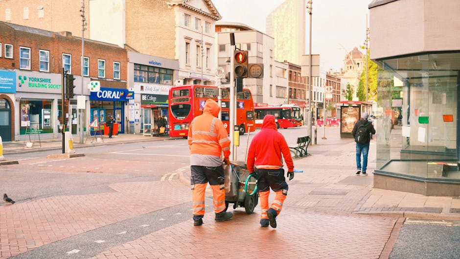 Two waste collection workers dressed in high-visibility orange trousers with reflective stripes and orange jackets are walking on a paved street in an urban area, pushing a green wheeled bin or trolley containing rubbish or equipment. They are positioned in the foreground, with their backs to the camera, moving towards a traffic light that is red. Behind them, the street features a variety of shops including a pharmacy, a building with a sign reading 'Coral', and other commercial premises, with some pedestrians walking nearby. Several red double-decker buses are visible in the background, indicating a busy city environment. The surrounding architecture includes mid-rise and taller buildings with a mix of modern and older facades, suggesting this is a commercial district with nearby offices or shops. The scene is illuminated by natural daylight, with soft light creating a slightly warm tone, and the area appears to be part of a private waste collection service, situating this scene within an alternative rubbish removal context.