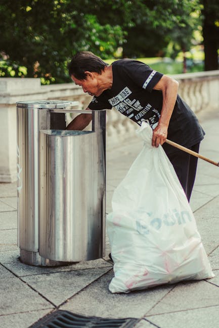 A collection of overflowing waste and rubbish bags situated on a paved sidewalk adjacent to a curb in an urban area. The scene includes black, red, and grey wheelie bins, with some lids open and debris spilling out, containing mixed waste such as cardboard boxes, plastic bags, paper, and packaging materials. Surrounding the bins are scattered boxes, paper, and discarded items, some flattened or torn, contributing to an unkempt appearance. Behind the rubbish, there are parked cars, including a silver hatchback, and a building with storefronts painted in yellow, green, and blue, featuring signage for local businesses. A metal railing separates the rubbish area from the parking and sidewalk, and a large tree trunk is visible on the left. The background features a multi-story building with scaffolding and safety barriers, indicating construction or refurbishment work nearby. The lighting suggests a cloudy day, emphasizing the cluttered, unmanaged waste scene typical of private rubbish collection in an urban environment.