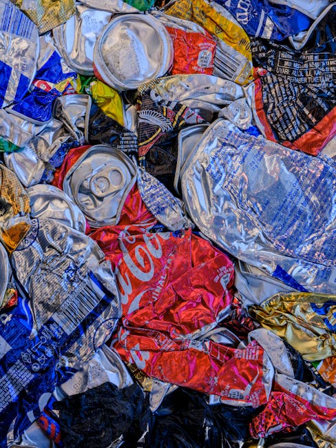 A close-up view of a large pile of crushed aluminium and plastic beverage cans, predominantly silver with some red and black branding, including partial Coca-Cola logos visible on a few cans. The cans are tightly compressed, creating a textured surface with visible ridges and dents, reflecting light unevenly. The pile is situated outdoors, likely on a paved or concrete surface, with some cans overlapping others, emphasizing their compacted state. The background appears blurred but suggests an environment related to waste collection or disposal, possibly near a recycling or rubbish removal service, such as those provided by House Clearance Enfield. The overall scene highlights the typical aftermath of recyclable beverage containers awaiting processing or disposal in an environmentally conscious waste management context.