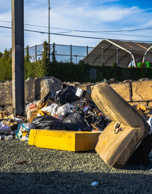 A waste collection vehicle is parked on a narrow urban street, actively collecting rubbish from a blue wheeled bin held by a worker dressed in a high-visibility orange vest and blue work clothes. The vehicle, primarily white with visible rust and dirt on its rear compartment, is positioned alongside a row of older, multi-story buildings with light-colored facades and large windows. The truck's rear loading area, which is open and appears to be designed for compacting and transporting waste, extends into the street, while the worker carefully places the bin into the vehicle’s opening. To the left, a black car is parked parallel to the curb, and a no-parking traffic sign is mounted on one of the buildings. The scene is lit by natural daylight, casting soft shadows on the cobblestone-paved street, illustrating a typical urban rubbish collection during the daytime, aligned with private waste handling and rubbish removal services often provided by companies such as House Clearance Enfield.