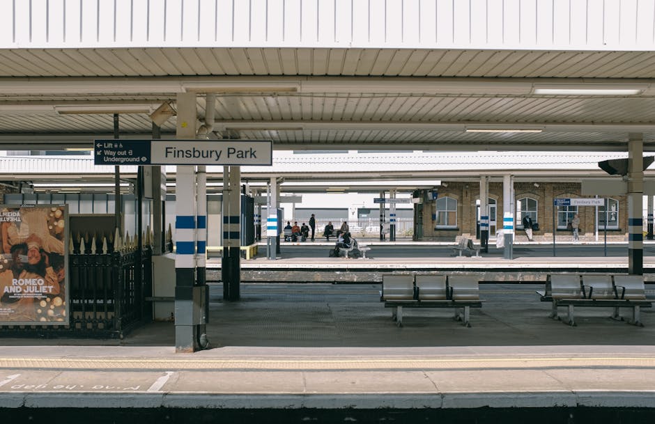The image depicts a view of Finsbury Park railway station platform, featuring a row of metal benches aligned parallel to the train tracks. The platform surface appears to be made of concrete with a tactile paving strip near the edge for safety. Overhead, a large, pale-colored corrugated metal roof provides shelter, supported by multiple vertical metal columns painted with blue and white stripes. In the background, several passengers are seated and standing, waiting for arriving or departing trains. To the left, a black metal fence and a lit poster advertising a theatrical production are visible, with the poster featuring an image of a woman and the title 'Romeo and Juliet.' The station environment is well-lit, with natural daylight filtering through gaps in the roofing, and environmentally, this scene represents an urban railway station setting. This imagery aligns with the context of transportation hubs, relevant for discussions about public transportation and alternative waste handling or rubbish disposal in busy communal spaces, as exemplified by services like House Clearance Enfield providing on-site or idle-time collection options in such environments.