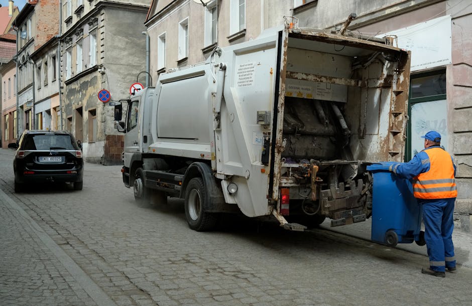 A street scene showing a white waste collection vehicle with an open rear compartment, positioned parallel to the pavement in an urban residential area. The truck's rear is rusted and contains empty mechanical components used for compacting or collecting rubbish. To the right, a worker wearing a high-visibility orange vest, blue overalls, and a blue cap is disposing of waste into a blue wheeled bin, which is being emptied into the truck. The street has cobblestone paving, and parked cars are visible along the curb, including a black vehicle with a European license plate. The buildings lining the street are multi-storey structures with light-colored facades, some showing signs of weathering or architectural details typical of older city areas. The scene is lit with natural daylight, with a generally neutral, professional tone suitable for an article about private rubbish collection or waste management services in urban environments, as provided by House Clearance Enfield.